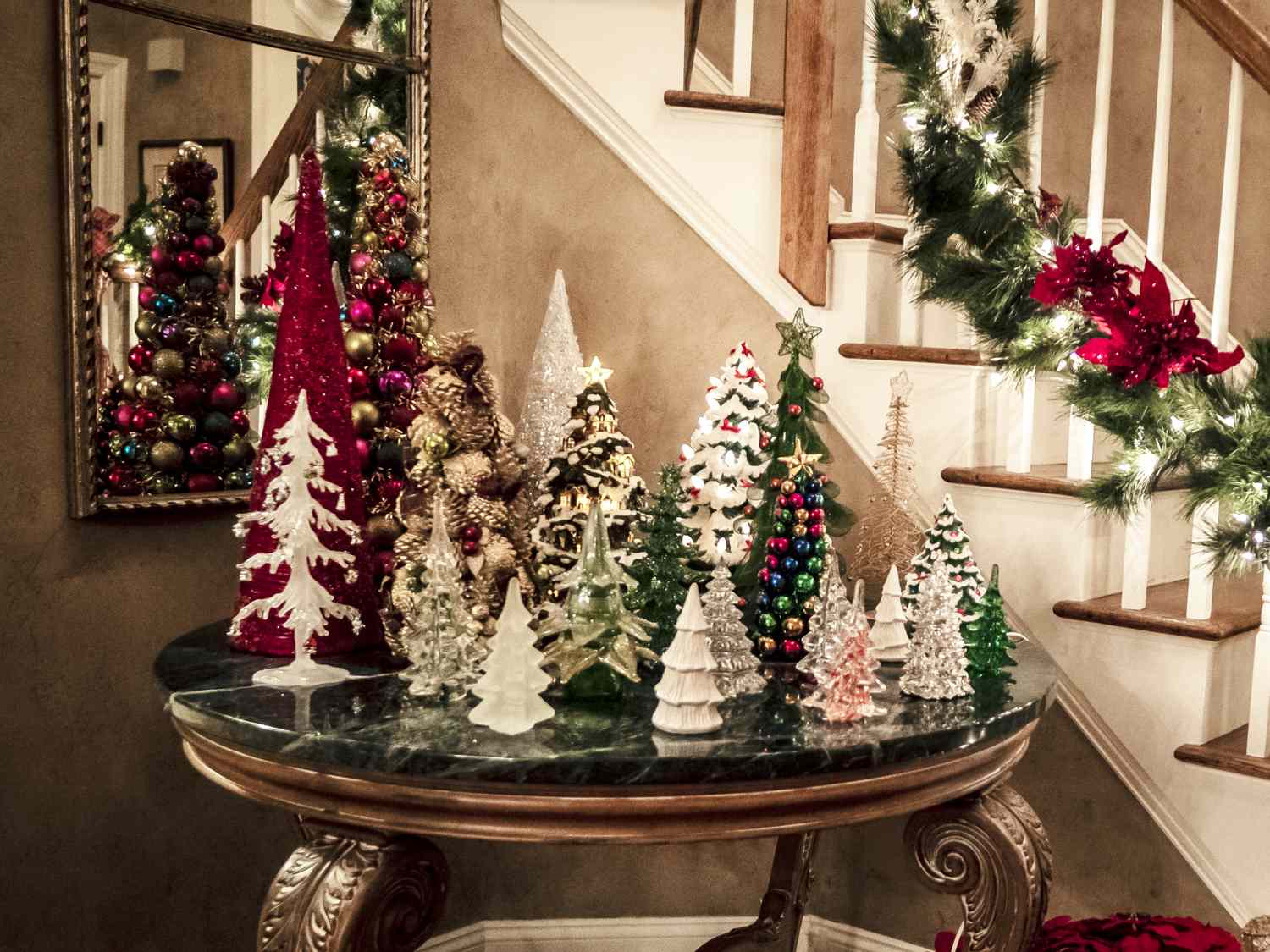 A table arranged with decorative Christmas trees and ornaments near a staircase decorated with holiday lights and garland