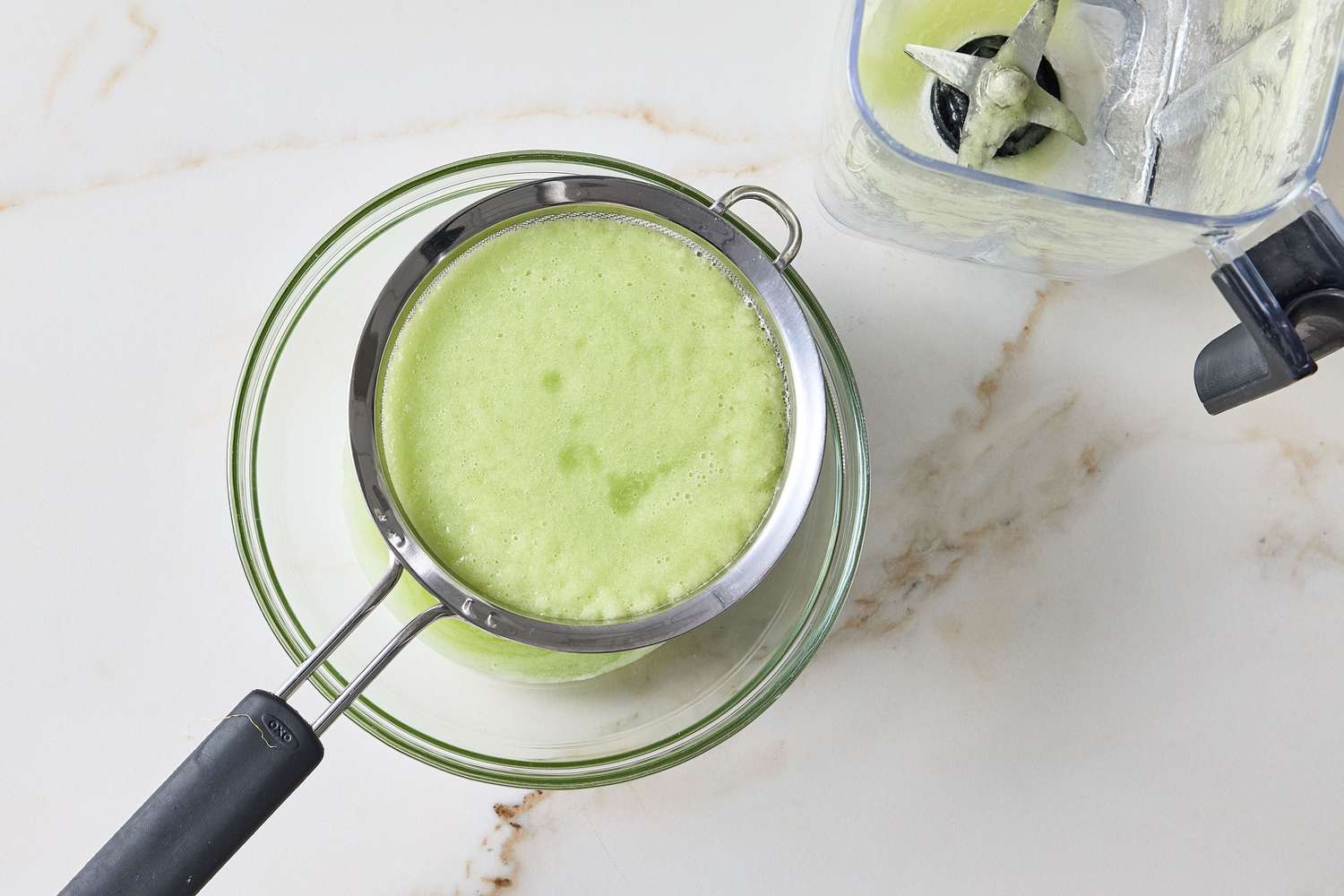 A green liquid being strained through a sieve into a glass bowl next to a blender container
