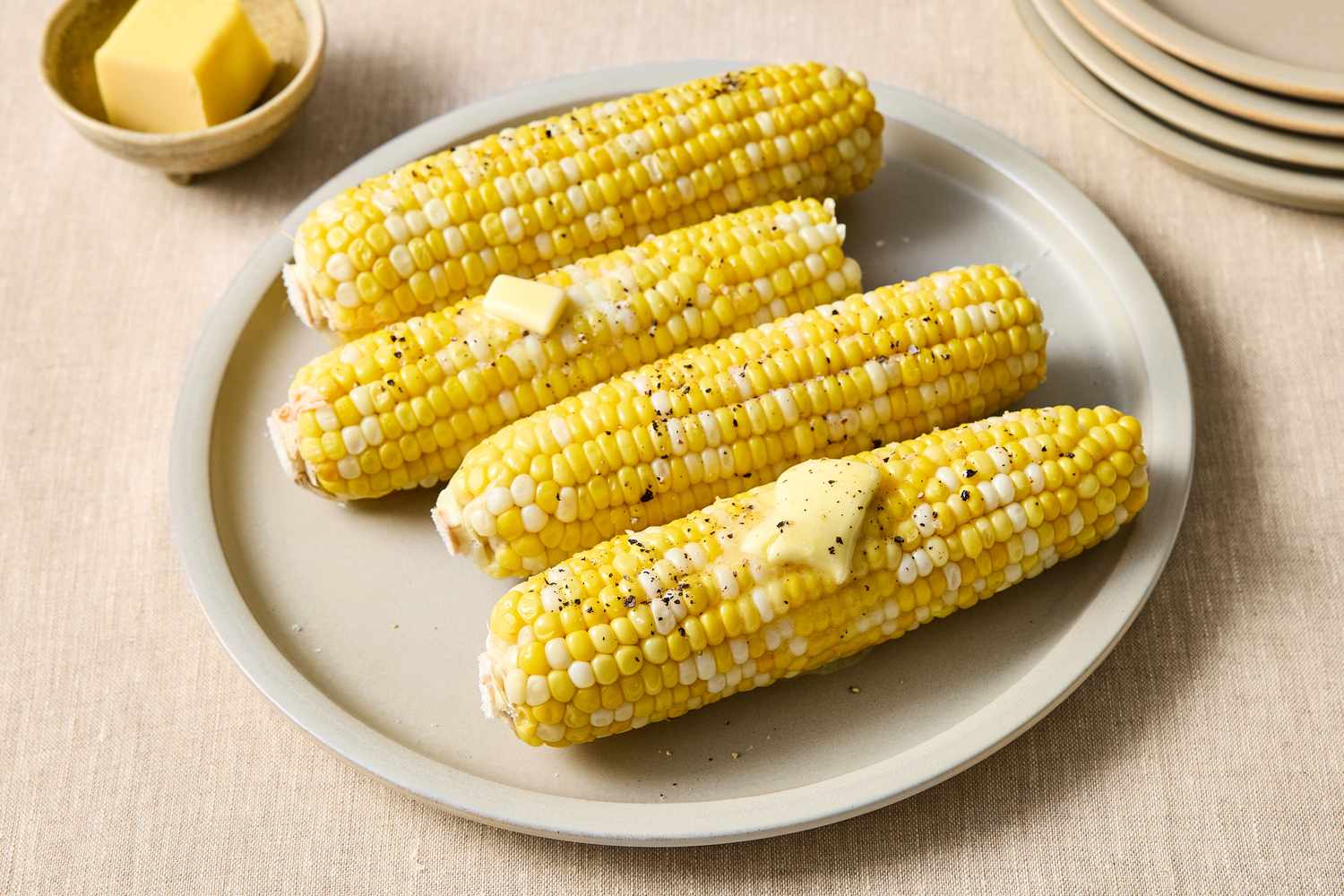 Four ears of corn on a plate topped with butter and ground pepper with a butter dish in the background