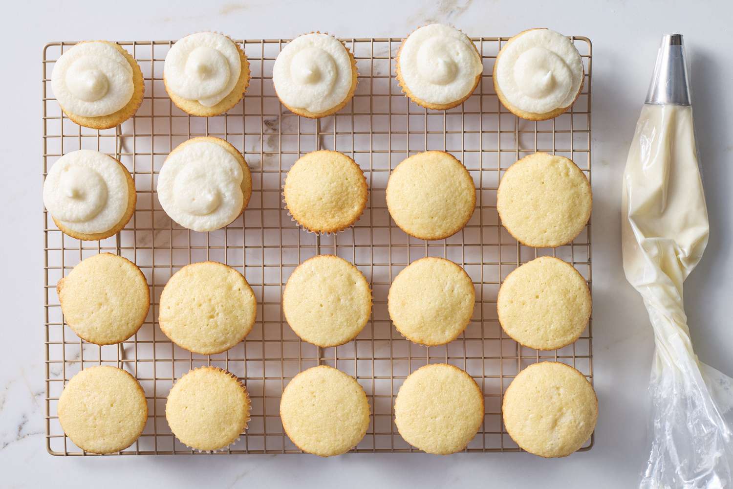 Tray of vanilla cupcakes some with frosting applied using a piping bag positioned on a cooling rack