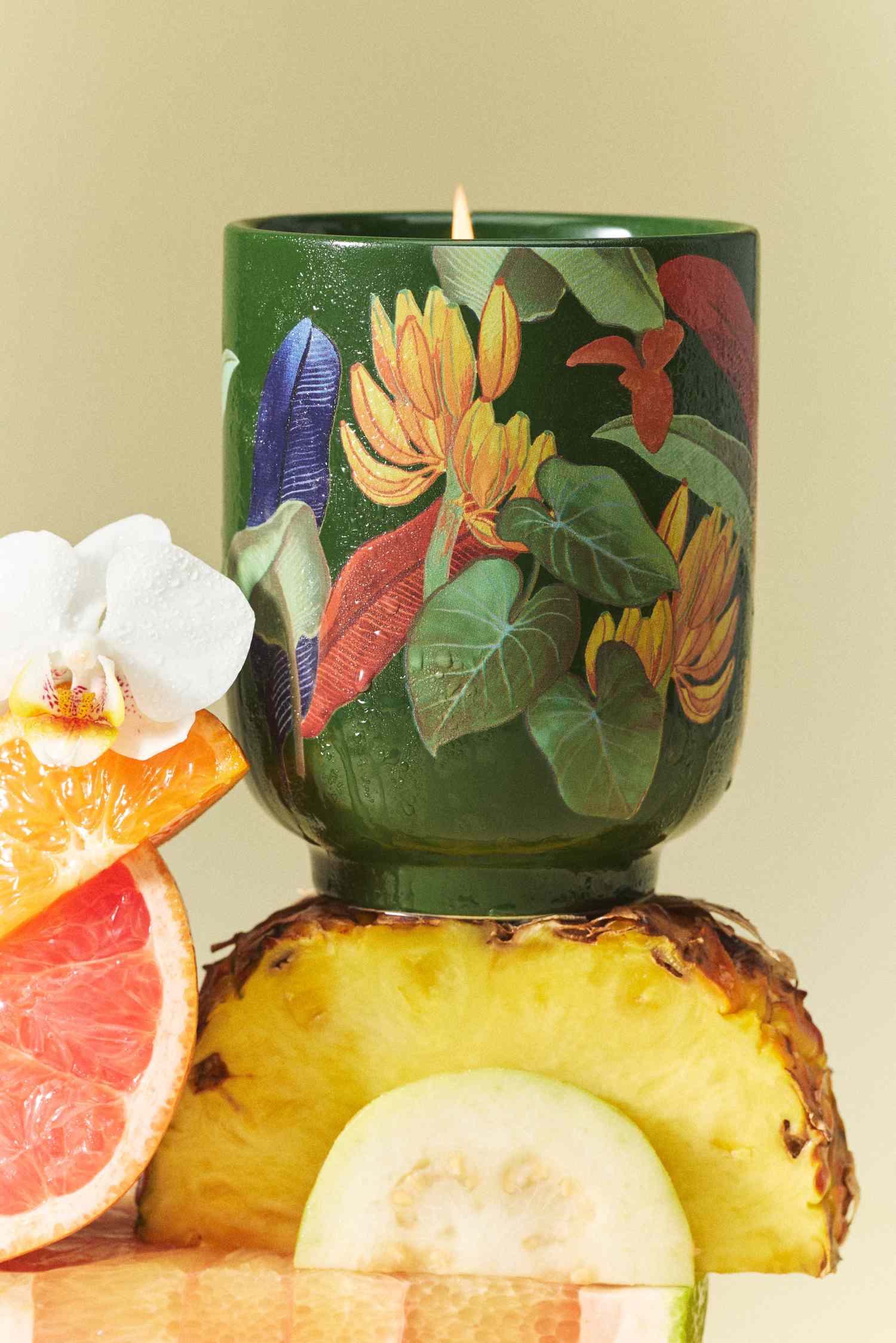 Decorative candle in a green holder with floral patterns displayed on tropical fruits