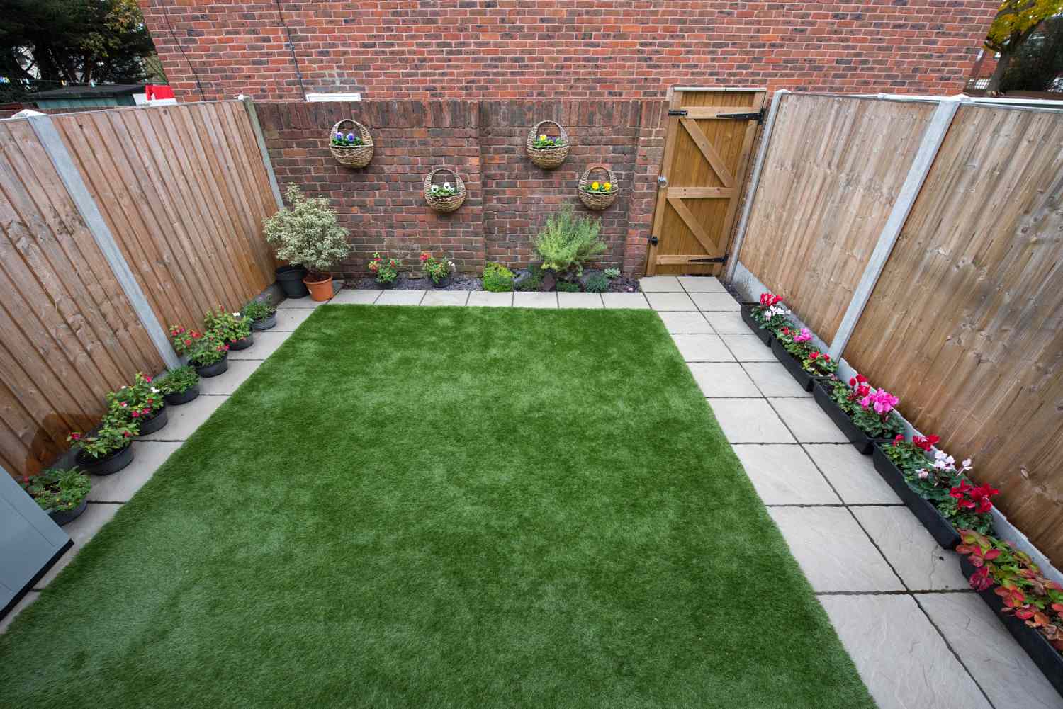Backyard with artificial turf surrounded by a paved path decorative plants on the wall and flowers along the edges