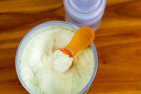 A tin of baby formula powder with a scoop on top a baby bottle in the background