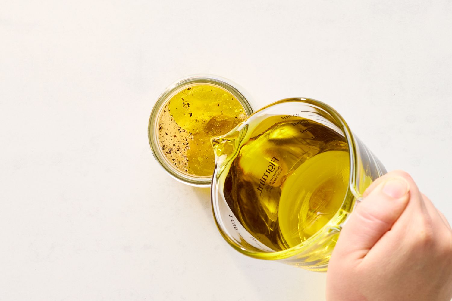 A hand pouring vinaigrette from a clear glass measuring cup into a small jar on a white surface