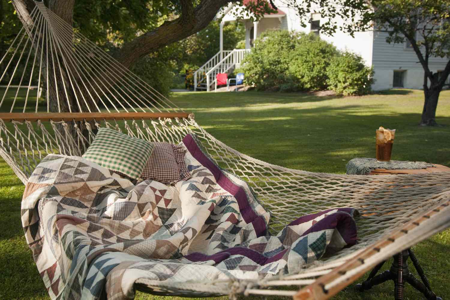 A colorful quilt and pillows on a hammock in a grassy backyard with a house in the background
