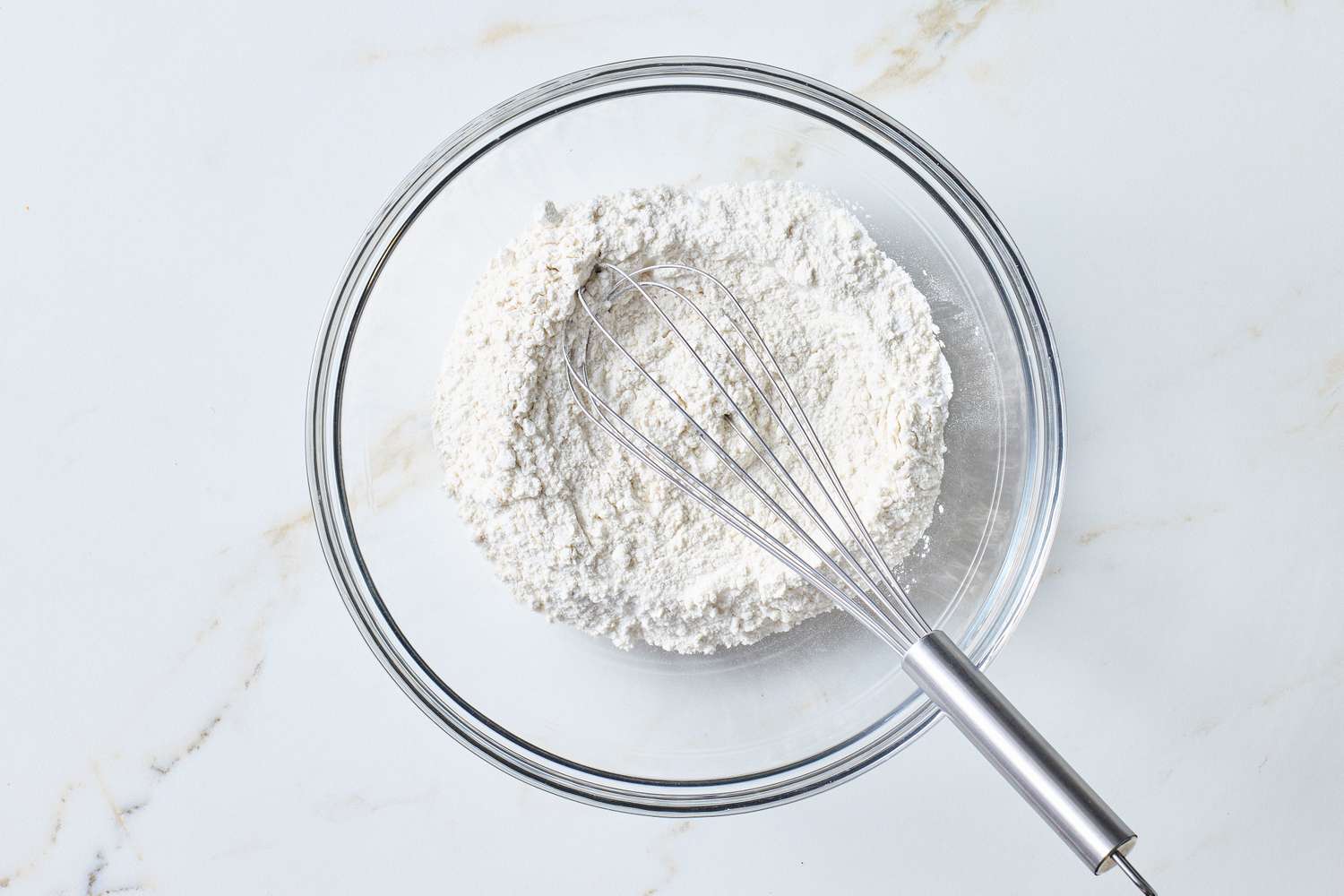 A mixing bowl filled with flour and a whisk on a marble countertop