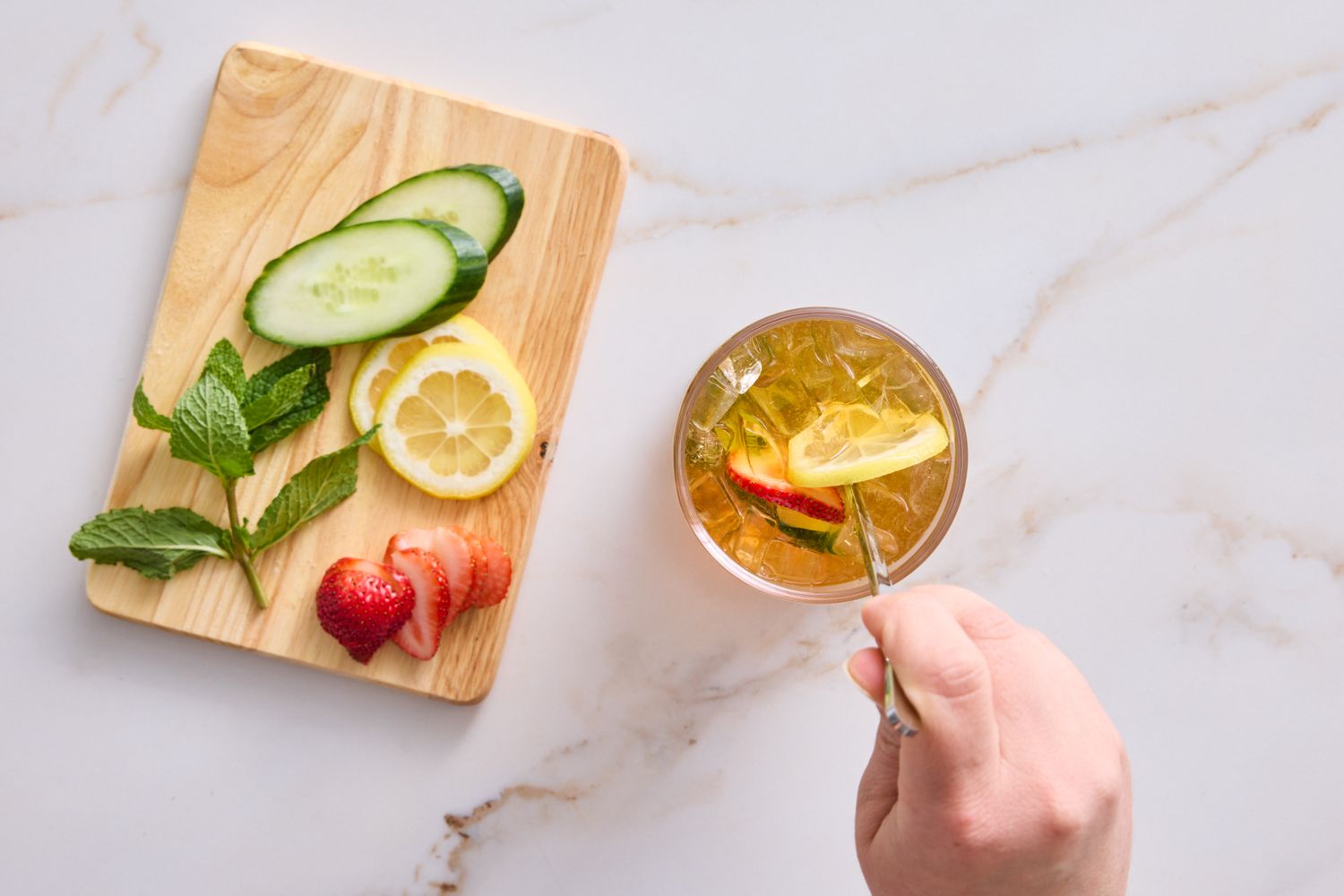 A hand stirring a drink with fruit garnish next to a wooden board with sliced garnish ingredients like cucumber lemon and mint