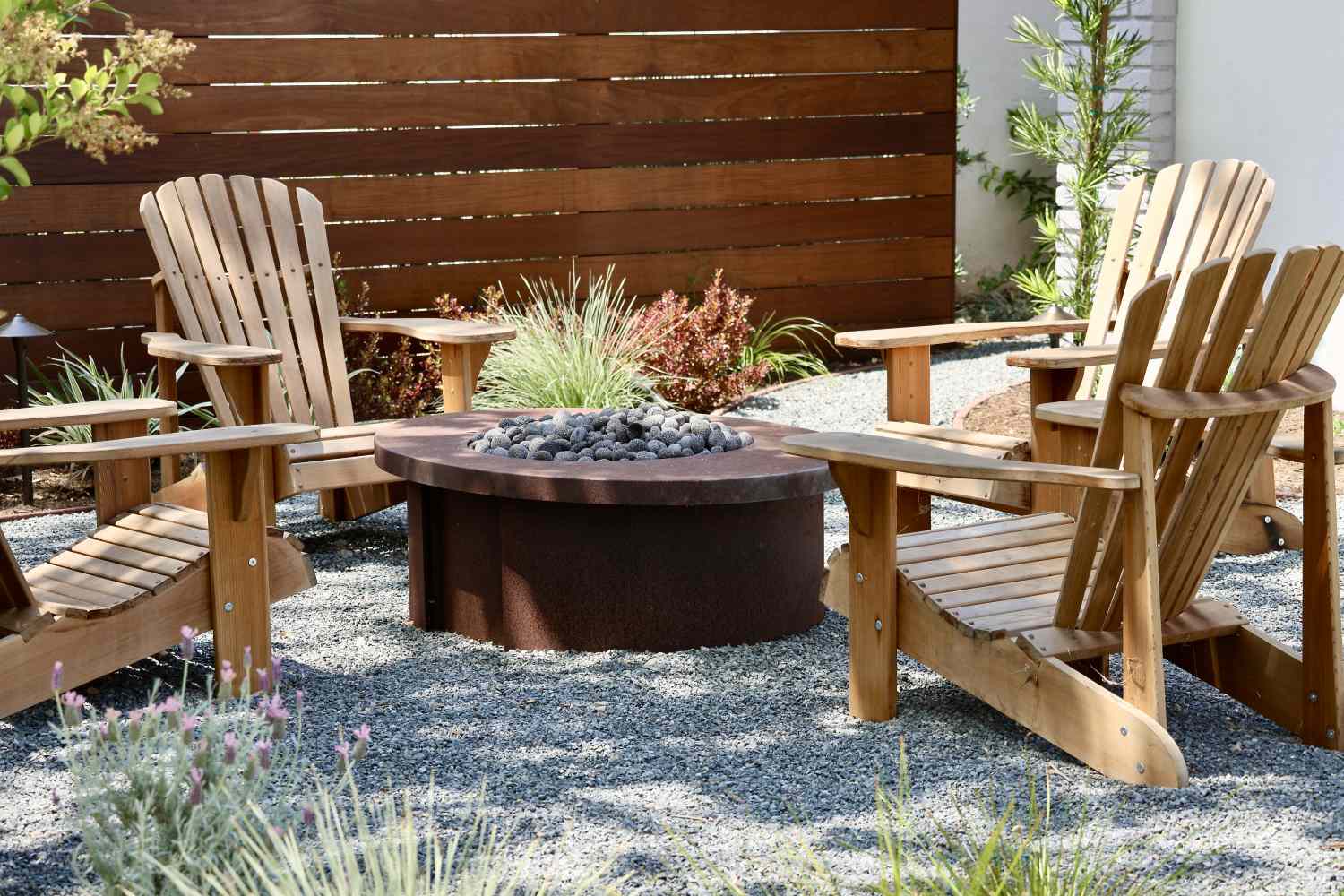 Four wooden outdoor chairs surrounding a fire pit with nearby plants and a wooden fence background