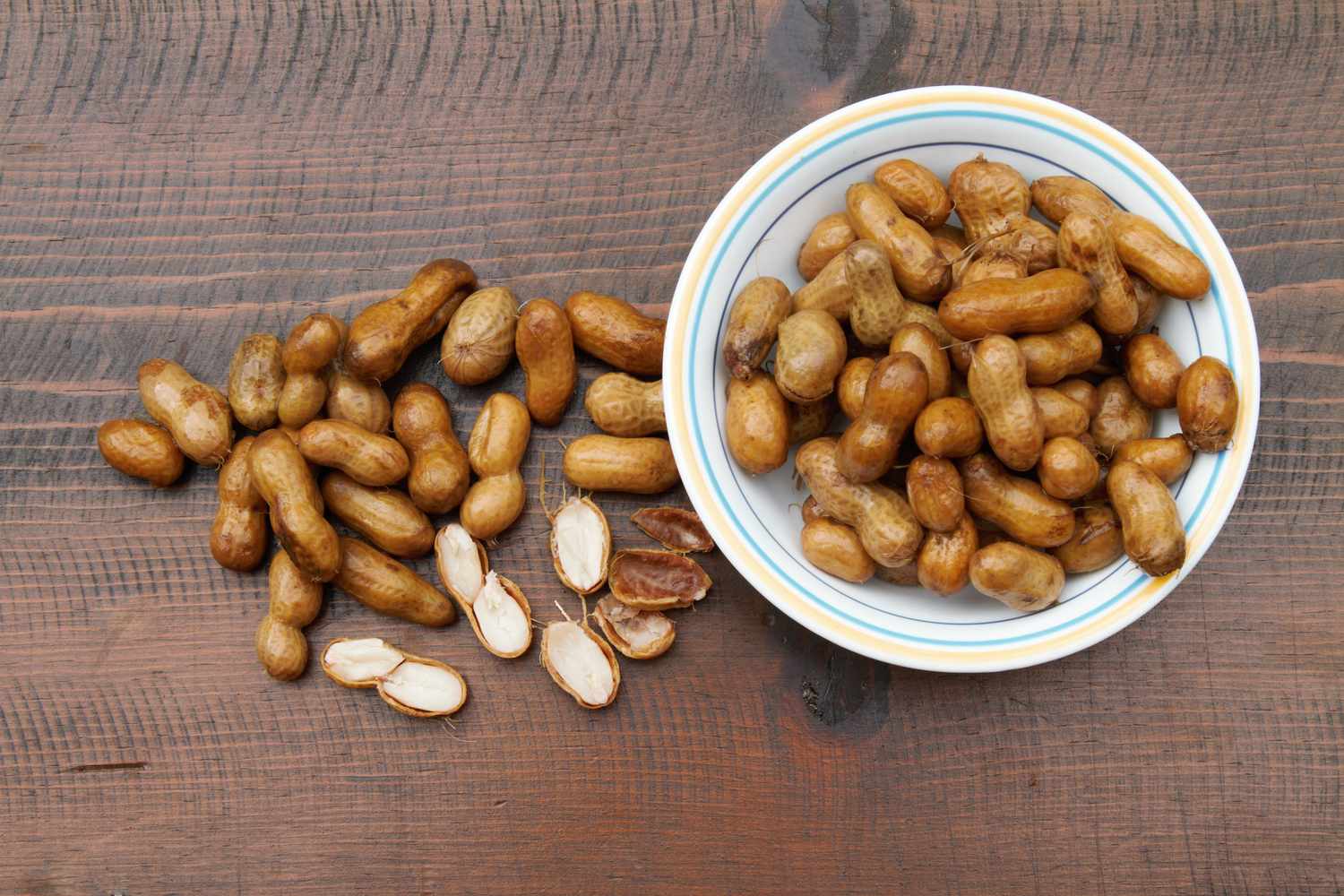 A bowl of boiled peanuts next to loose peanuts on a wooden surface with some peanuts split open showing their insides