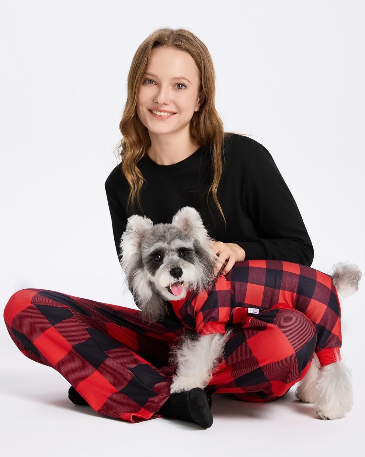A person sits on the floor holding a small dog wearing a matching plaid pattern outfit