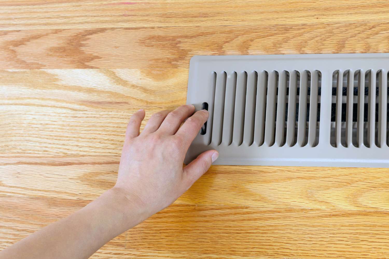 A hand adjusting a vent on a wooden floor