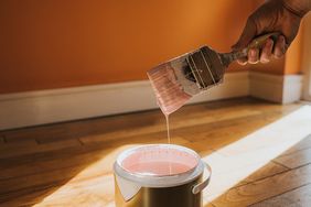 A hand holding a paintbrush with paint dripping into a can in a room with a wooden floor