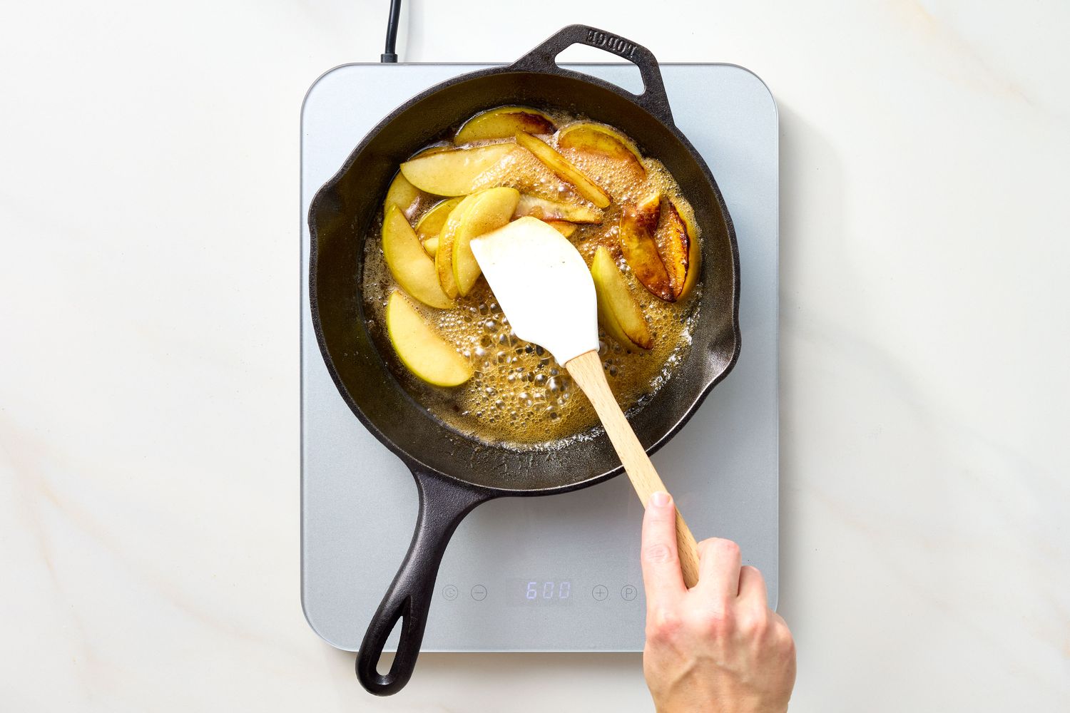 A persons hand cooking sliced apples in a cast iron skillet on an electric stovetop
