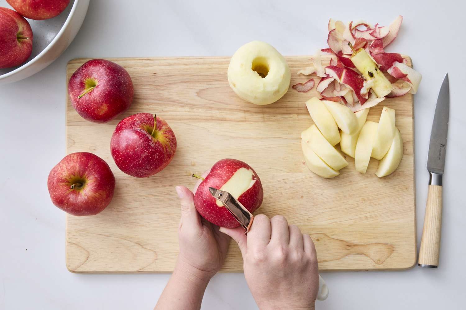 A person peeling apples on a wooden cutting board surrounded by peeled sliced and whole apples