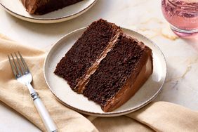 A slice of chocolate layer cake with icing on a plate accompanied by a fork and beige napkin