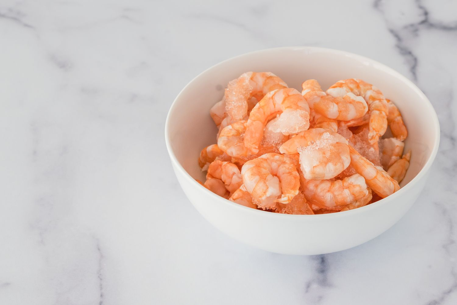 A white bowl filled with cooked shrimp placed on a marble surface