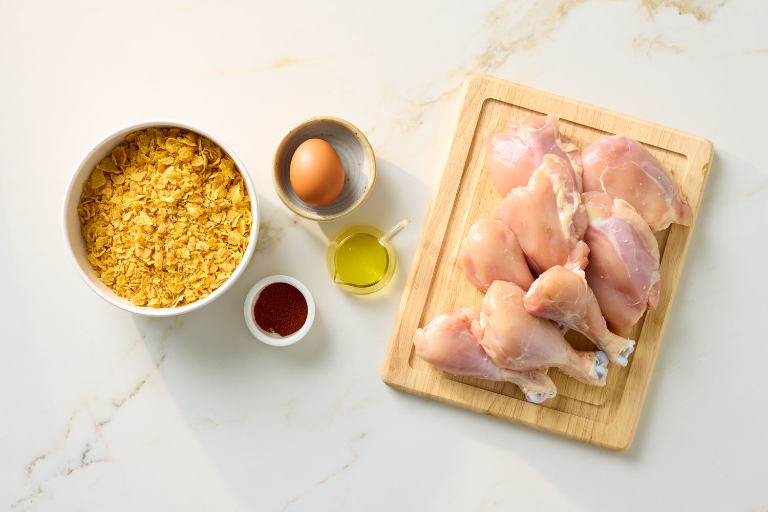 Chicken drumsticks and ingredients including cornflakes an egg olive oil and paprika on a white surface