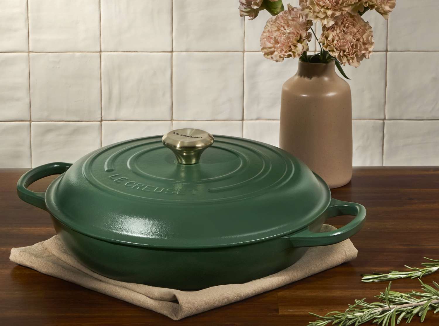 A green braiser with a lid on a wooden countertop near a vase with flowers and a sprig of rosemary