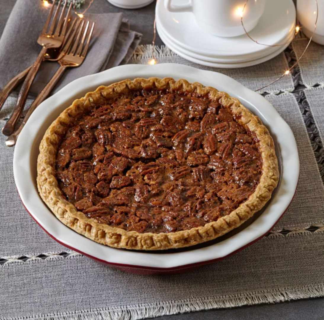 A pecan pie in a white dish set on a table with utensils and teacups nearby