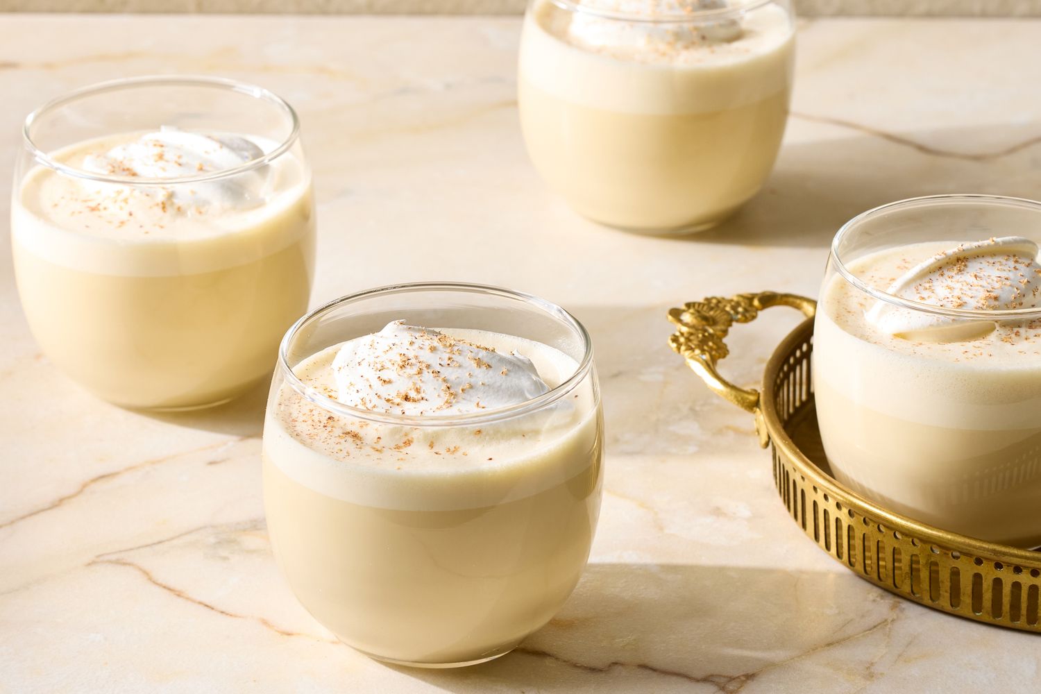 Four glass cups of eggnog with whipped cream on top placed on a creamcolored surface with one in a decorative holder