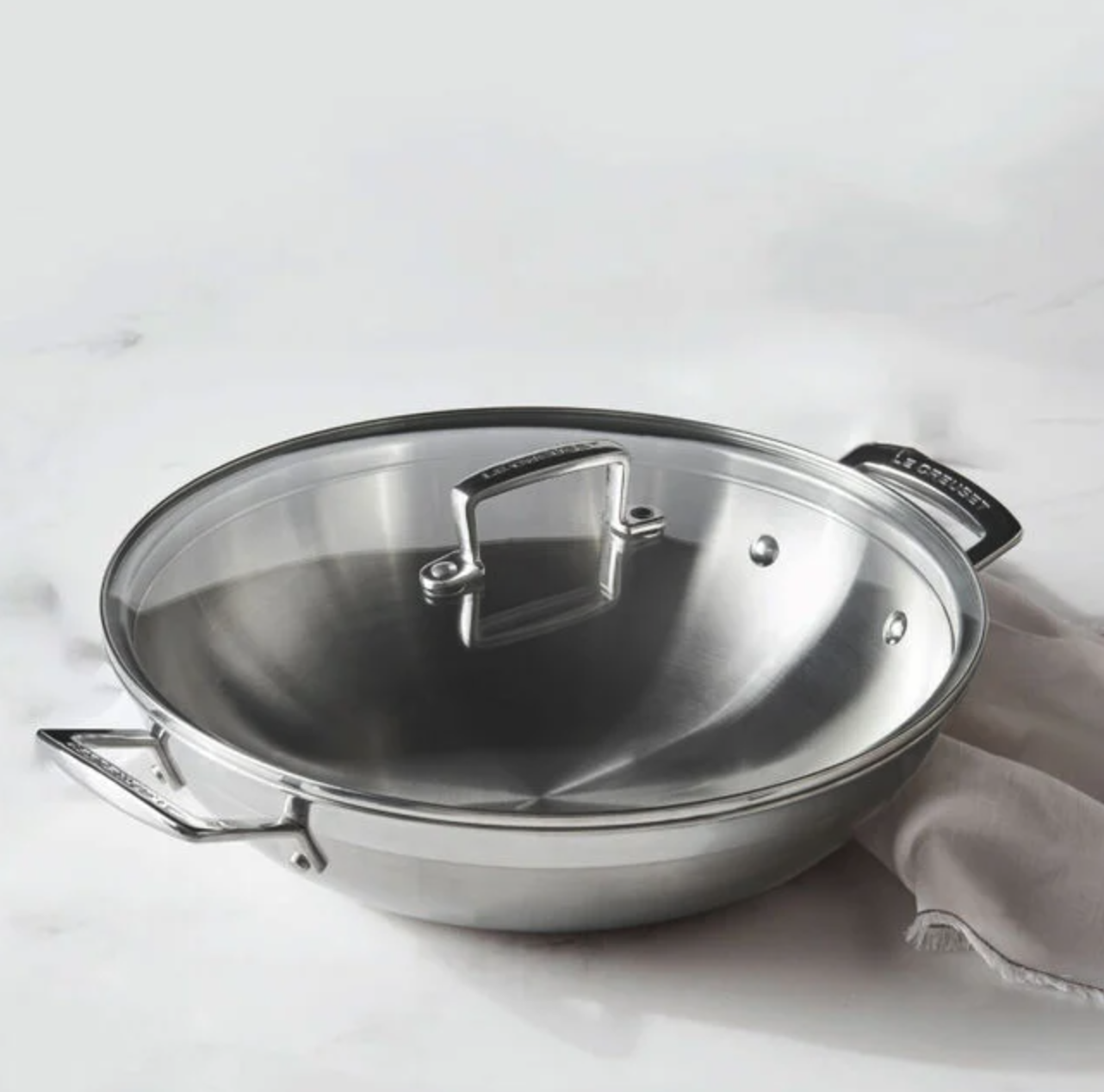 A stainless steel wok with a glass lid and cloth next to it