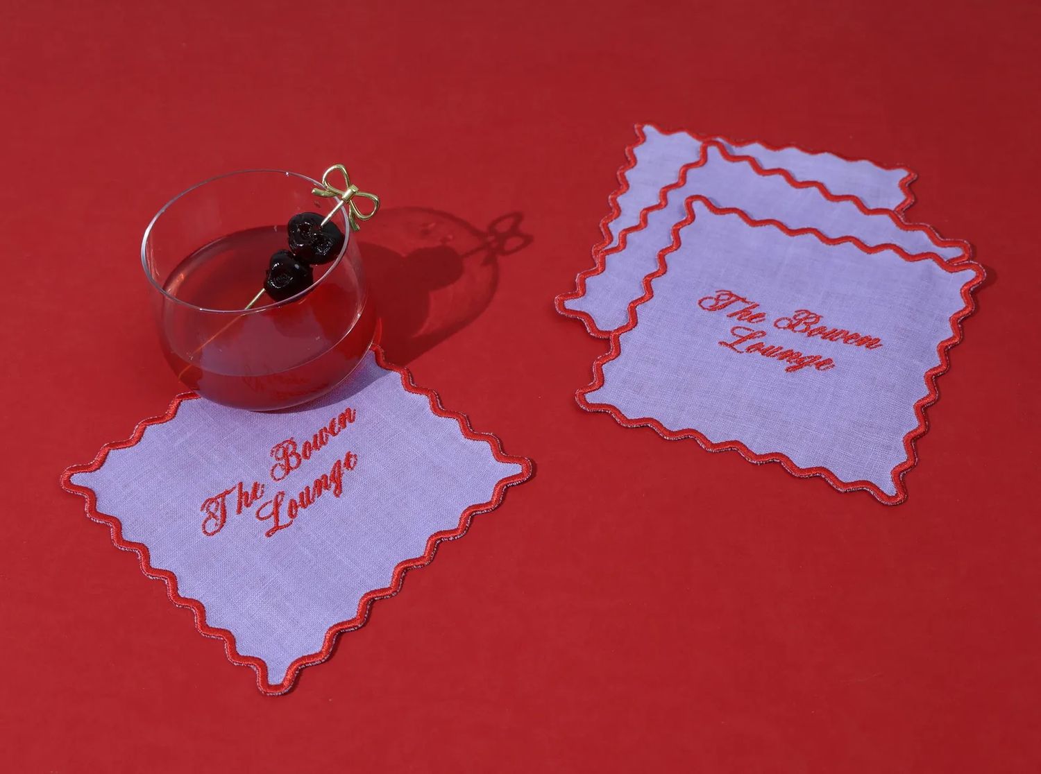 A cocktail glass with cherries on a napkin embroidered with The Brown Lounge set against a red background with matching additional napkins