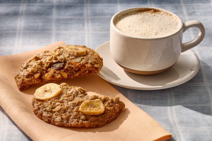 Two breakfast cookies with banana slices on a napkin beside a cup of coffee on a table
