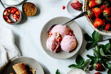 Bowl of strawberry ice cream with a spoon surrounded by strawberries and toppings on a white surface