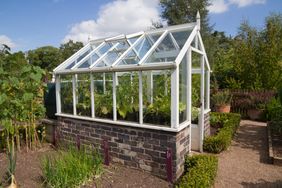 A greenhouse with plants inside situated in a garden environment with surrounding vegetation and a brick base