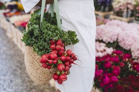 Close-up of a bag filled with fresh kale and radishes, held at an outdoor farmers market with flowers in the background