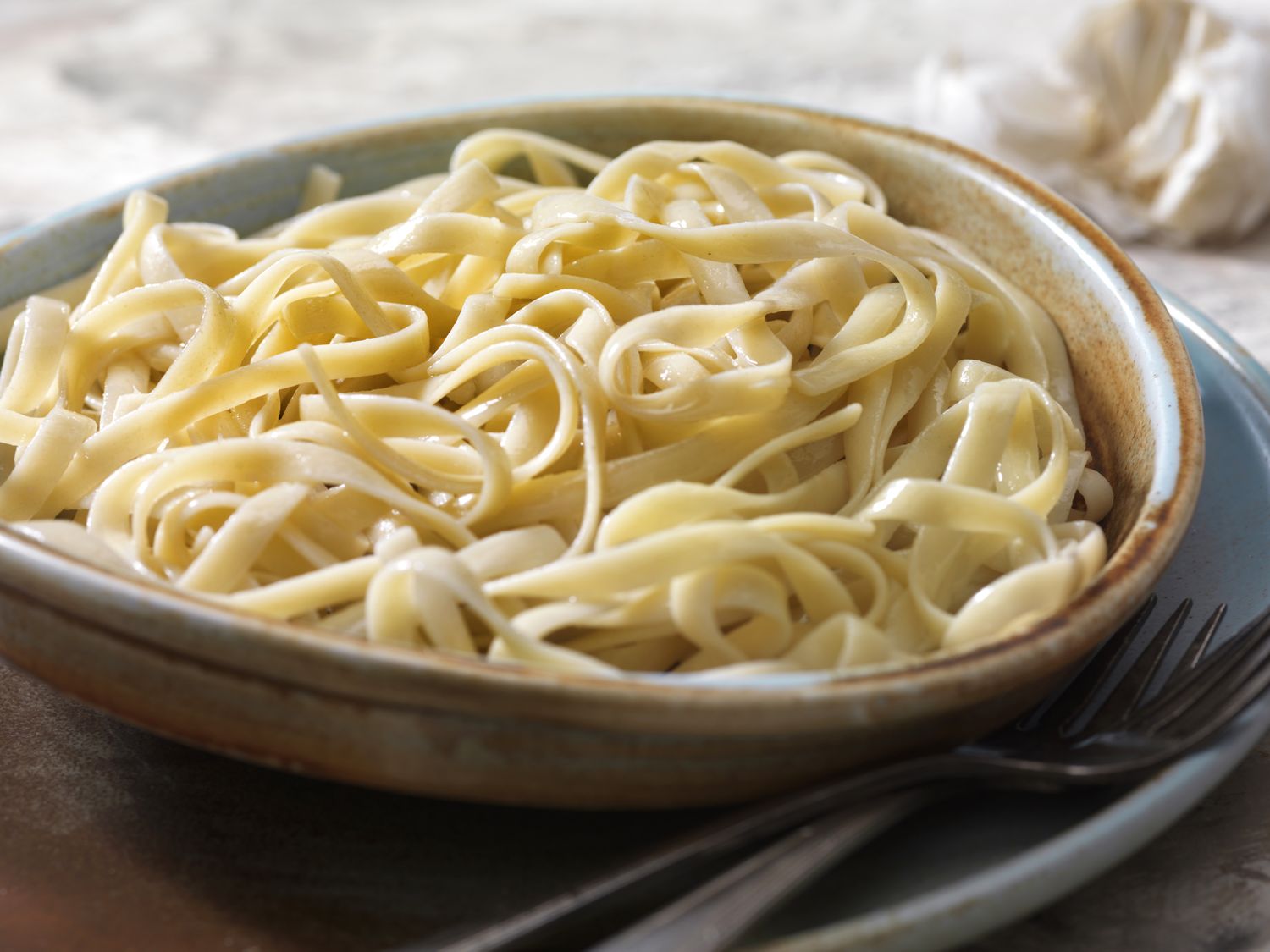 A bowl of fettuccine pasta with garlic clove in the background
