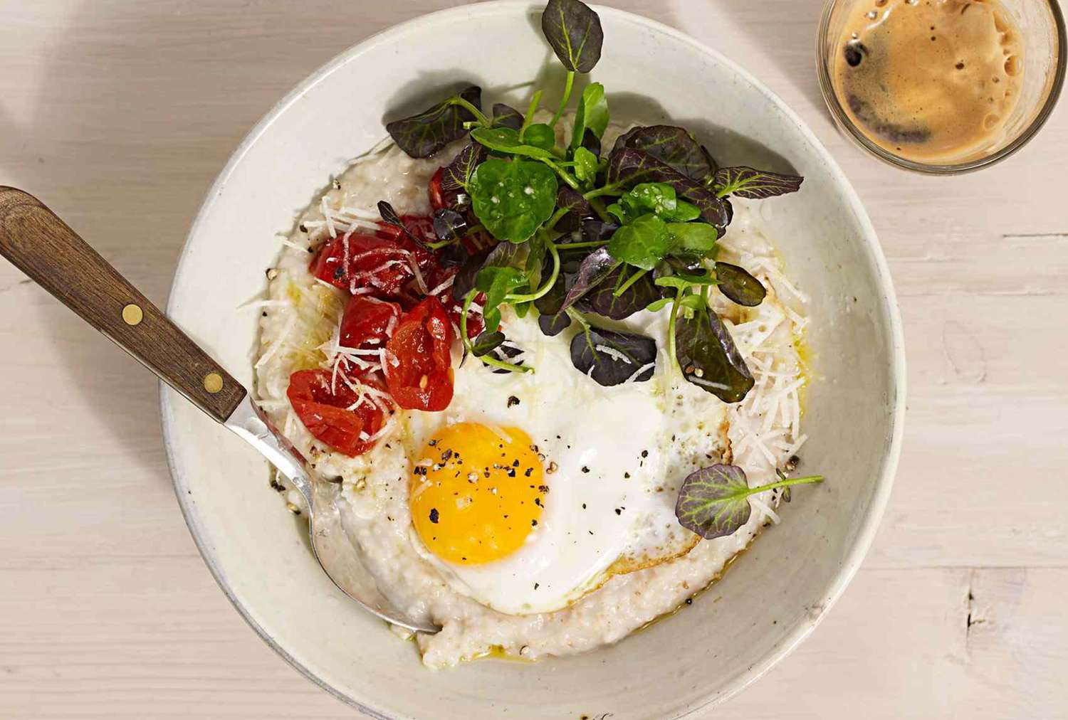 A bowl of savory oatmeal topped with a fried egg greens and cherry tomatoes accompanied by a cup of coffee