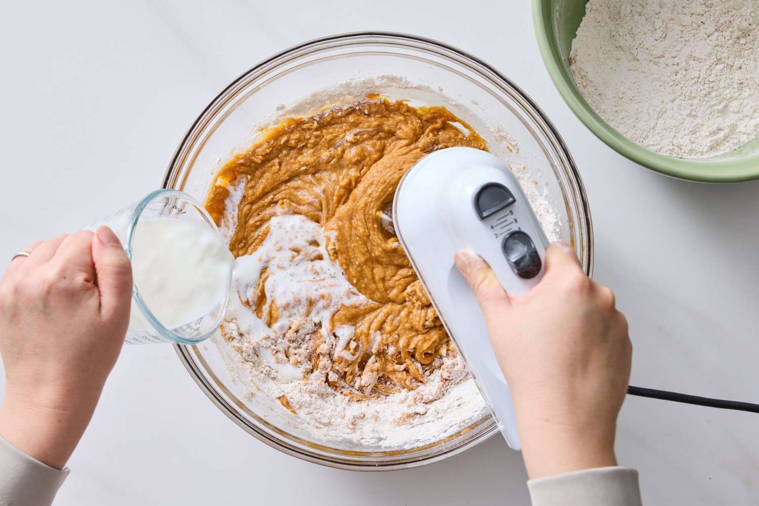 Hands preparing batter in a bowl with a mixer and pouring liquid from a cup
