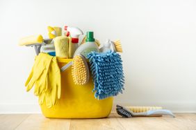 A bucket filled with various cleaning supplies and tools, such as gloves, brushes, and cloth, placed on a wooden floor