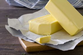 Two blocks of butter cut with a knife. Unwrapped and placed on a wooden chopping board, on a rustic wooden table.