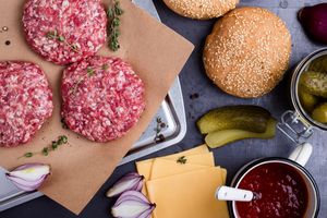 Raw hamburger patties on parchment paper beside burger ingredients like buns cheese slices pickles onions and ketchup