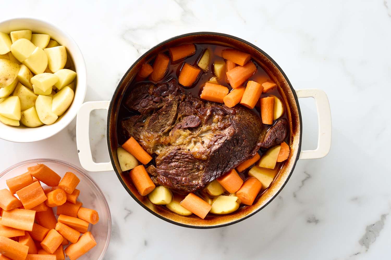 Pot roast in a casserole dish with carrots and potatoes surrounded by bowls of cut carrots and potatoes