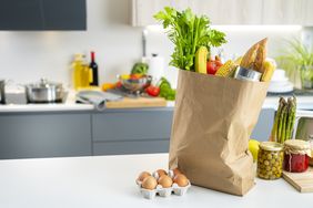A paper grocery bag filled with fresh produce and items placed on a kitchen counter next to a carton of eggs and jars of preserves