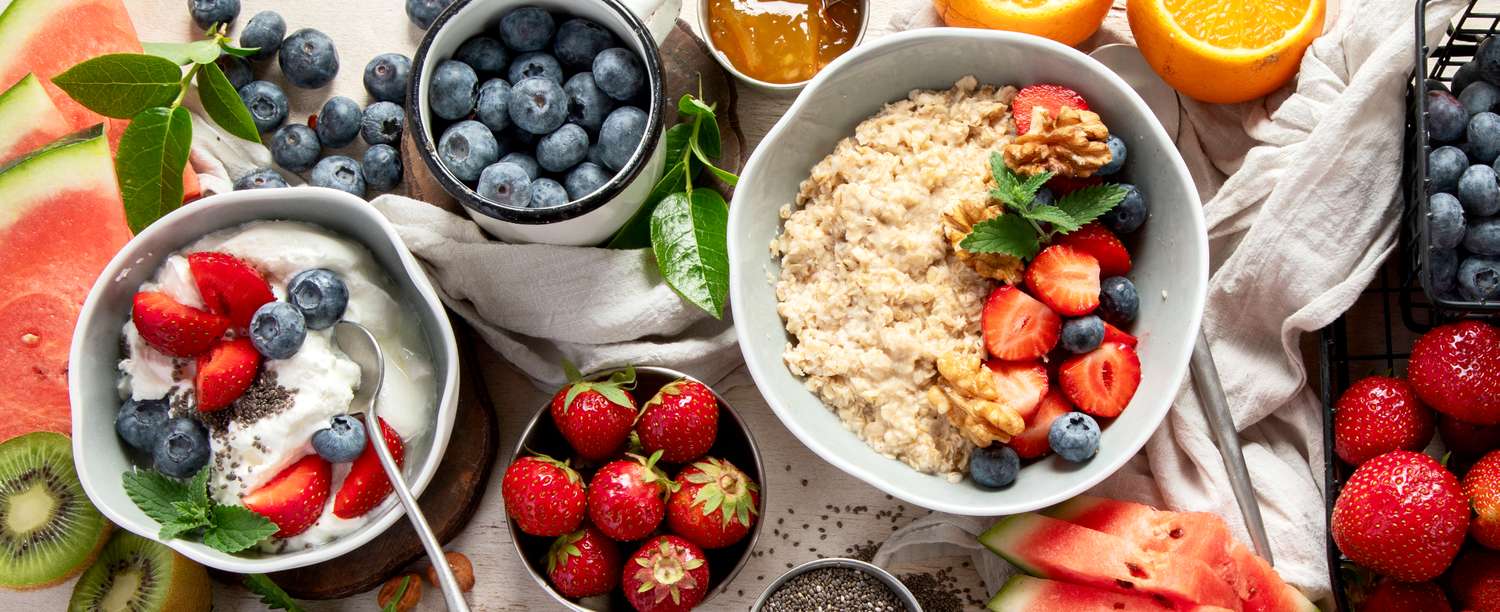 Various fruits and breakfast items including bowls of oatmeal and yogurt topped with fresh berries and slices of watermelon and kiwi