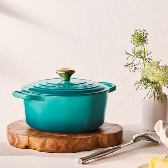 A lidded cooking pot sitting on a wooden board surrounded by a spoon flowers and a butter pat