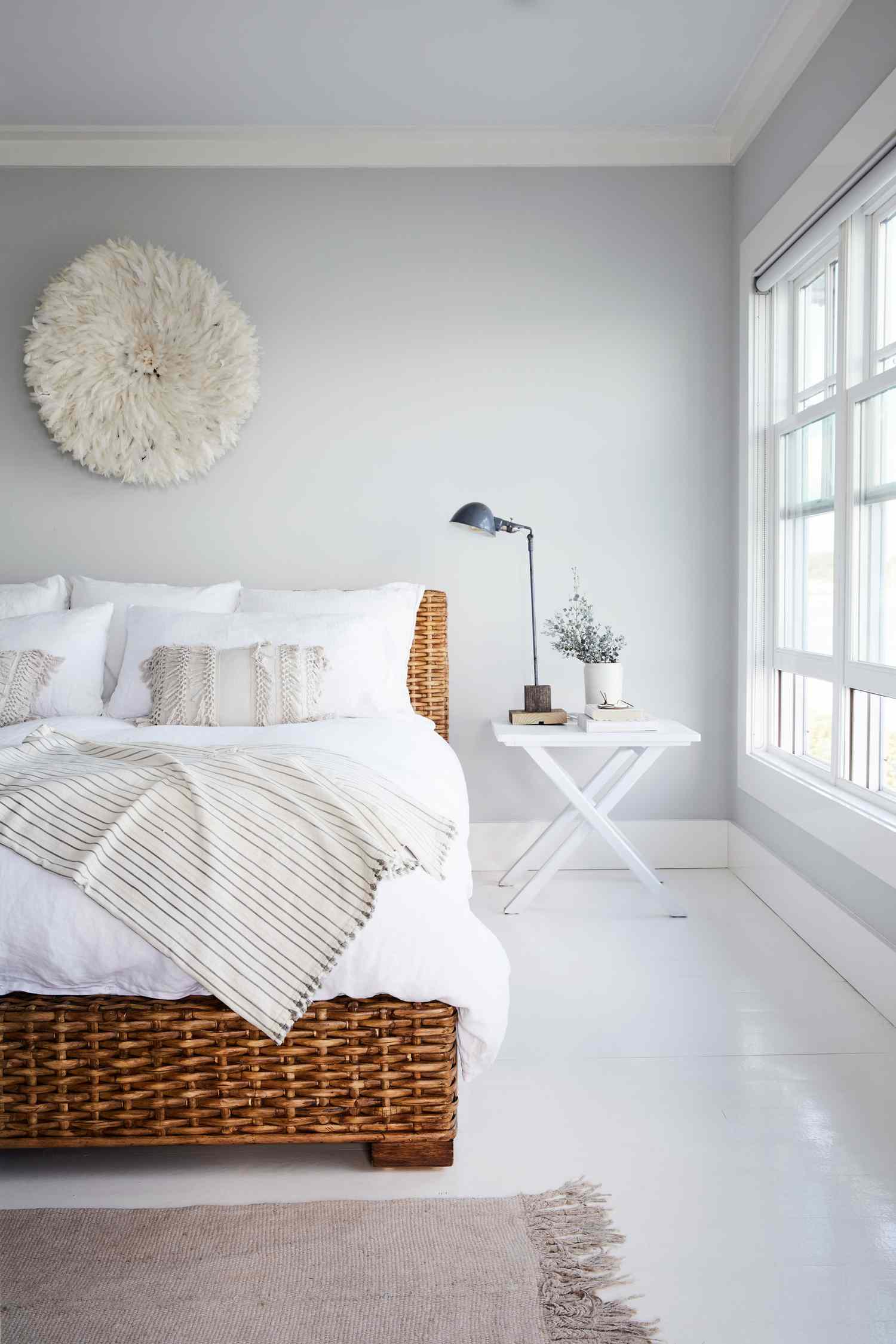 A bedroom with a wicker bed frame white bedding wall dcor and a side table near a window