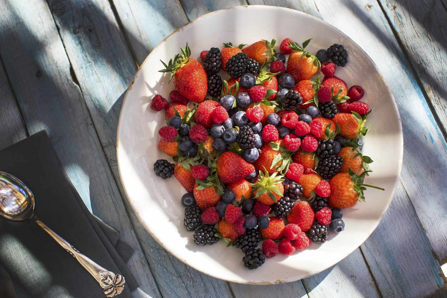 fresh strawberries, blueberries, raspberries, and blackberries on a white plate on a painted wooden surface
