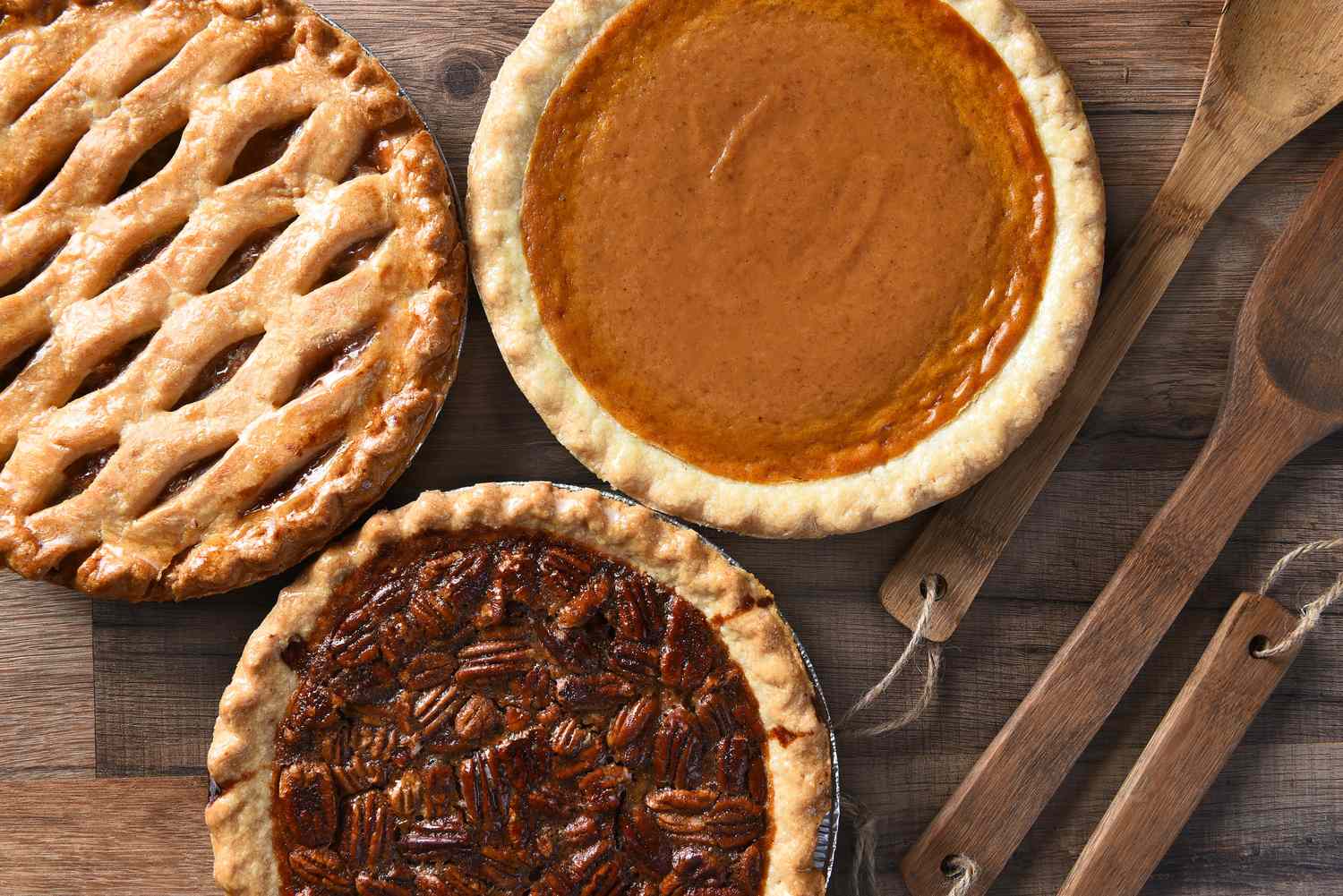 Three pies on a table one with a lattice crust one is pumpkin and one is pecan next to wooden serving utensils