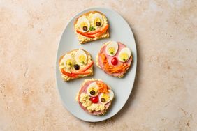 Four openface sandwiches on a plate decorated to resemble faces using eggs vegetables and deli meats