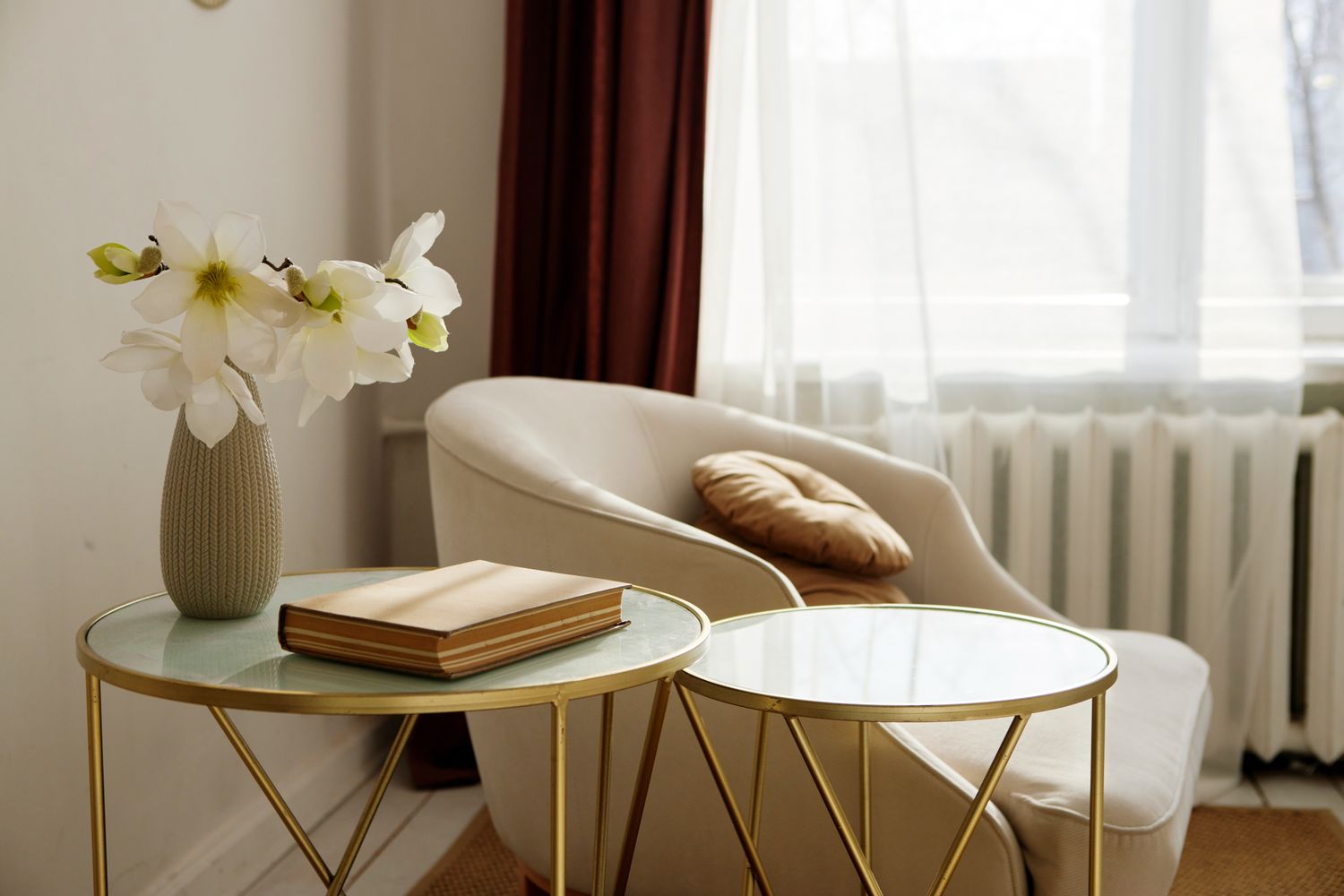 A cozy living space featuring a chair with a pillow a vase of flowers and two small tables with a book and glass tops