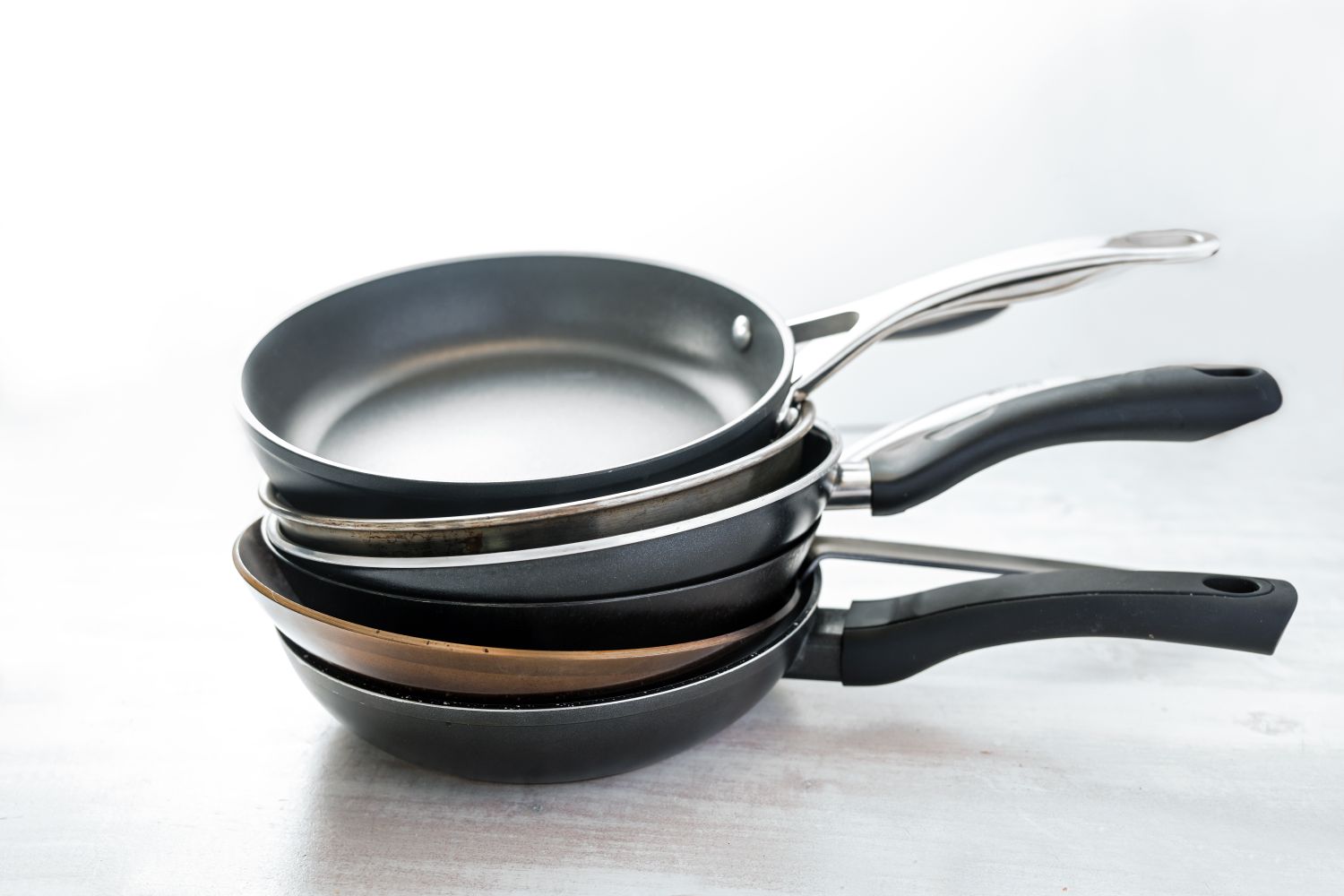 Stack of various frying pans made from cast iron, carbon steel, non-stick coated and stainless steel, choosing the right kitchen ware, light gray background, copy space, selected focus