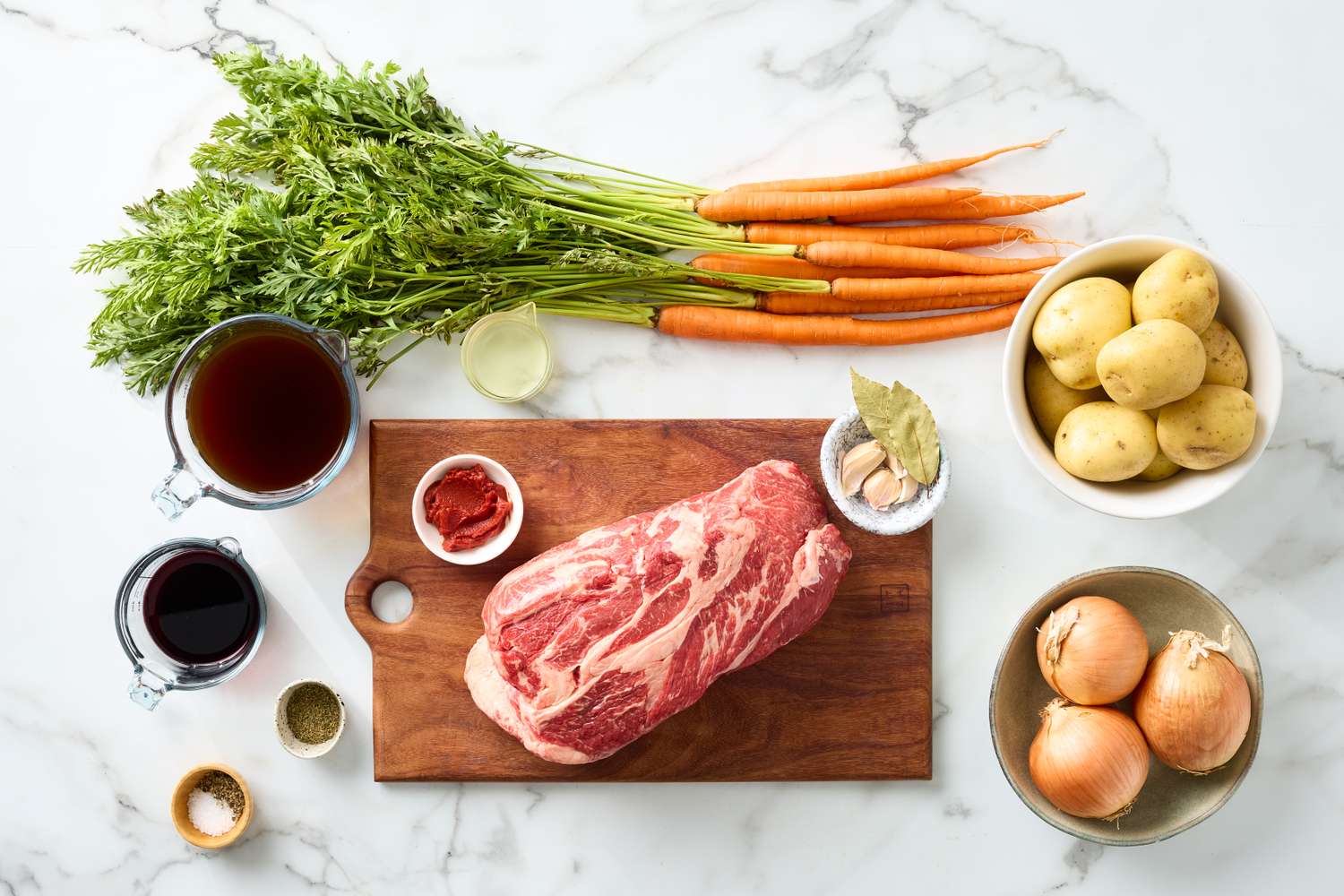 Ingredients for pot roast arranged on a surface including a large cut of meat carrots potatoes onions and other items