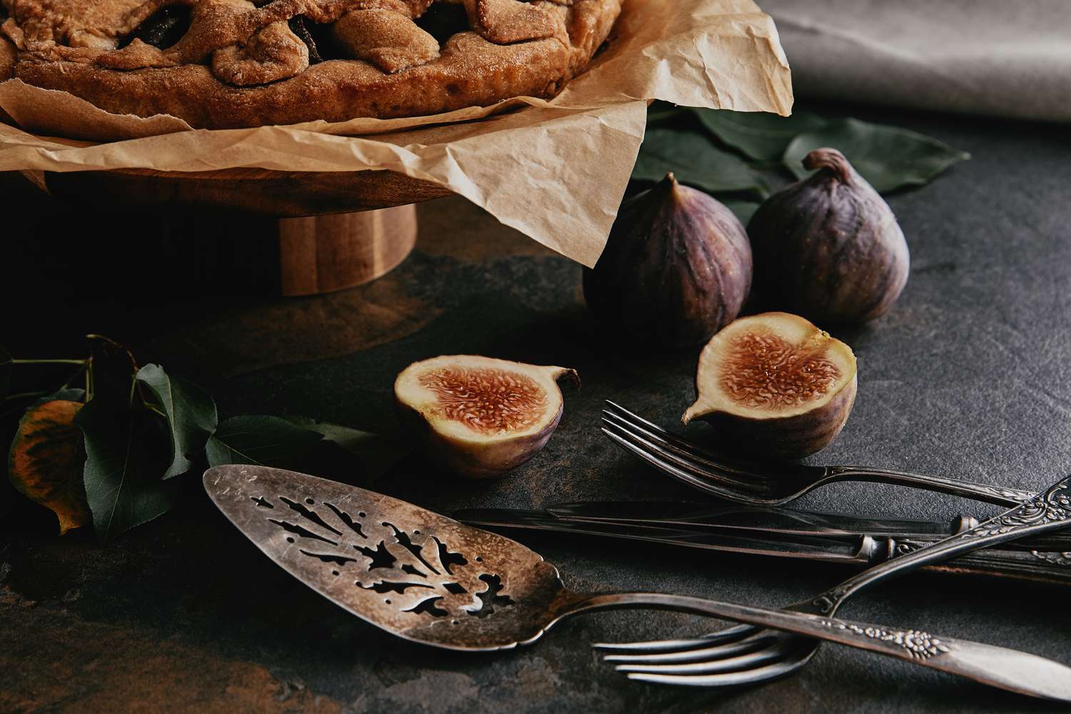 A pie on parchment paper with figs cutlery and a decorative pie server nearby