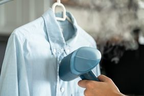A hand using a steamer on a shirt hanging on a hanger steam visible near the shirt