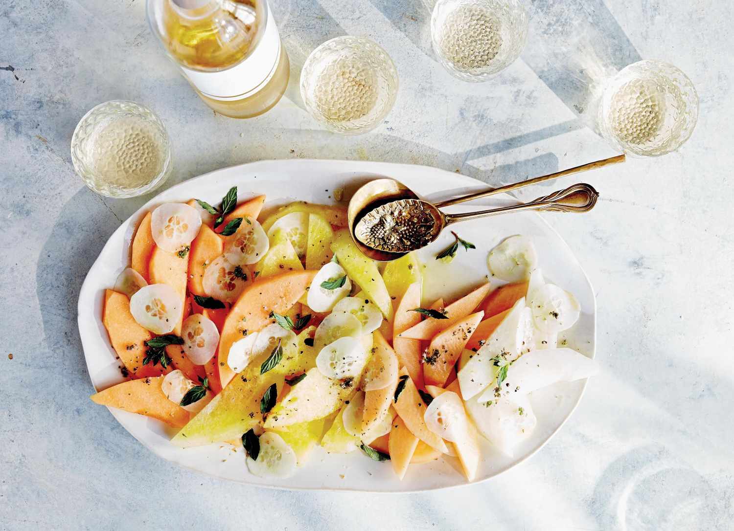 A plate of melon and cucumber salad with seasoning and a serving spoon glassware and a bottle in the background